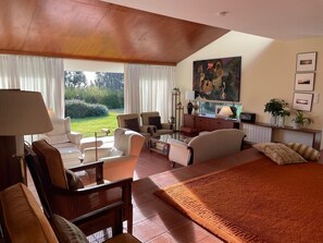 Amplia habitación con una cama grande, un sofá, una mesa de comedor y un espacio de escritorio. Se aprecia vista de vegetación a través de las ventanas.