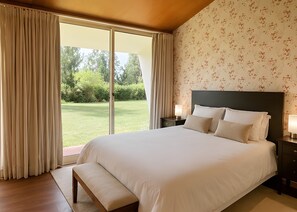 Un dormitorio con una cama grande, un banco, una mesita de noche con lámpara y vistas a un jardín a través de una puerta corredera.