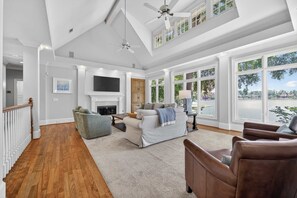 A spacious living room with a fireplace, large windows, and a high ceiling with a fan. - Sandy Cove Cottage- Great Waters Lakefront Luxury (Eatonton)