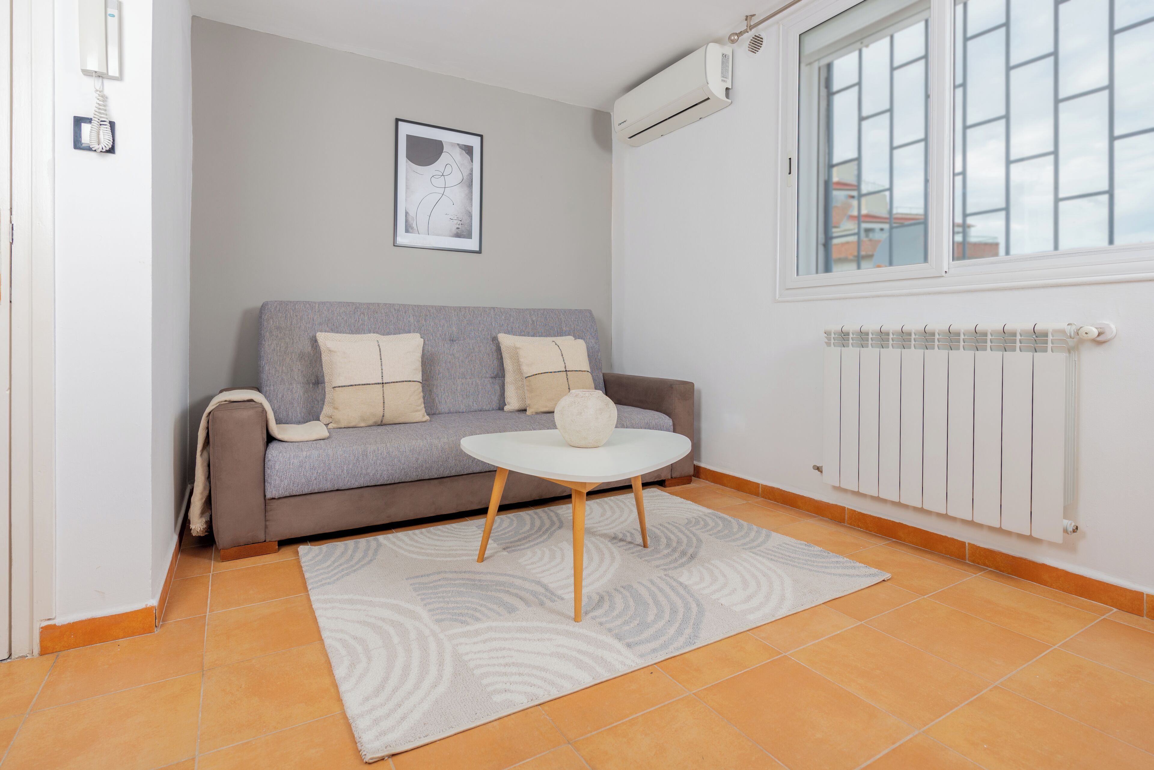 Una sala de estar con un sofá gris, una mesa de centro redonda blanca, un tapete gris estampado, una obra de arte abstracto enmarcada y una ventana con vista a edificios.
