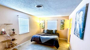 Une chambre à coucher avec un lit, deux fenêtres équipées de stores, une bouche d’aération au plafond, une étagère fixée au mur avec une plante et un tableau.