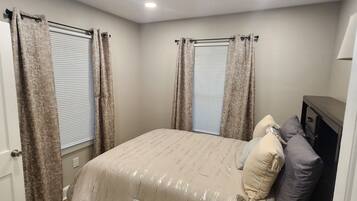 Une chambre à coucher avec un lit, des rideaux, une fenêtre avec des stores, une porte et un luminaire au plafond.