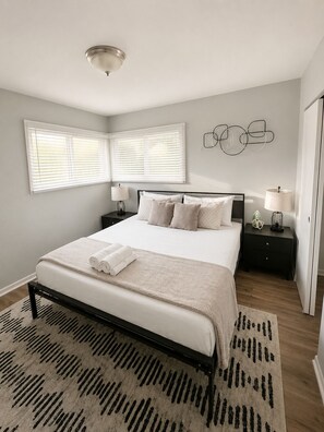 A bedroom with a bed, two bedside tables, a wall-mounted light fixture, and a window with blinds.