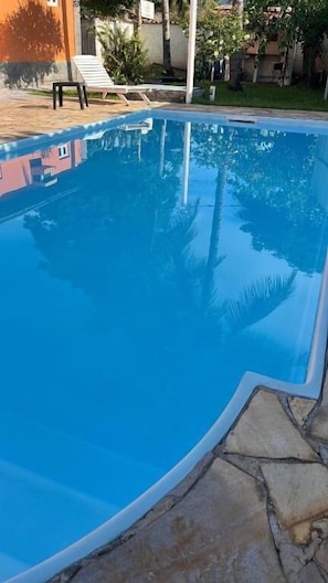 Una piscina con borde de piedra, rodeada de césped y algunos árboles.