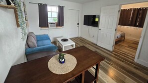 Un espace de vie compact comprenant un canapé, une petite table, un téléviseur fixé au mur et une porte donnant sur une autre pièce.
