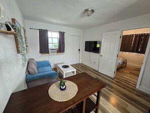 A compact living space with a sofa, a small table, a TV mounted on the wall, and a door leading to another room. - Cozy Place Near the Design District | Free parking (Miami)