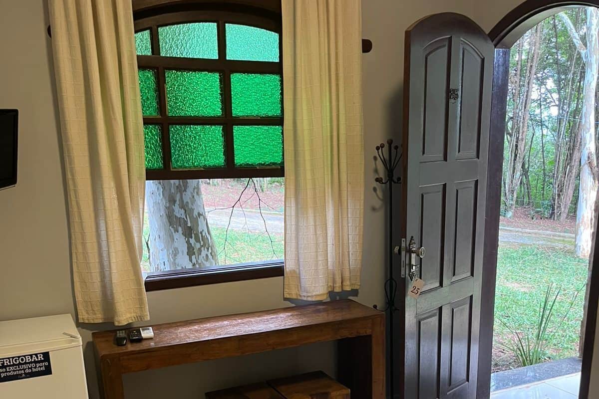Una habitación con una cama, un escritorio con una silla, un taburete pequeño, un refrigerador y una puerta que da al exterior.