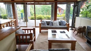 Ein Zimmer mit Esstisch aus Holz, einer Couch mit Kissen, einem Fernseher und Blick ins Freie.
