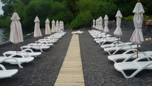 Weiße Liegestühle und Sonnenschirme reihen sich an einem Kiesweg, der zu einem Gewässer führt, umgeben von viel Grün.
