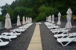 Weiße Liegestühle und Sonnenschirme reihen sich an einem Kiesweg, der zu einem Gewässer führt, umgeben von viel Grün.