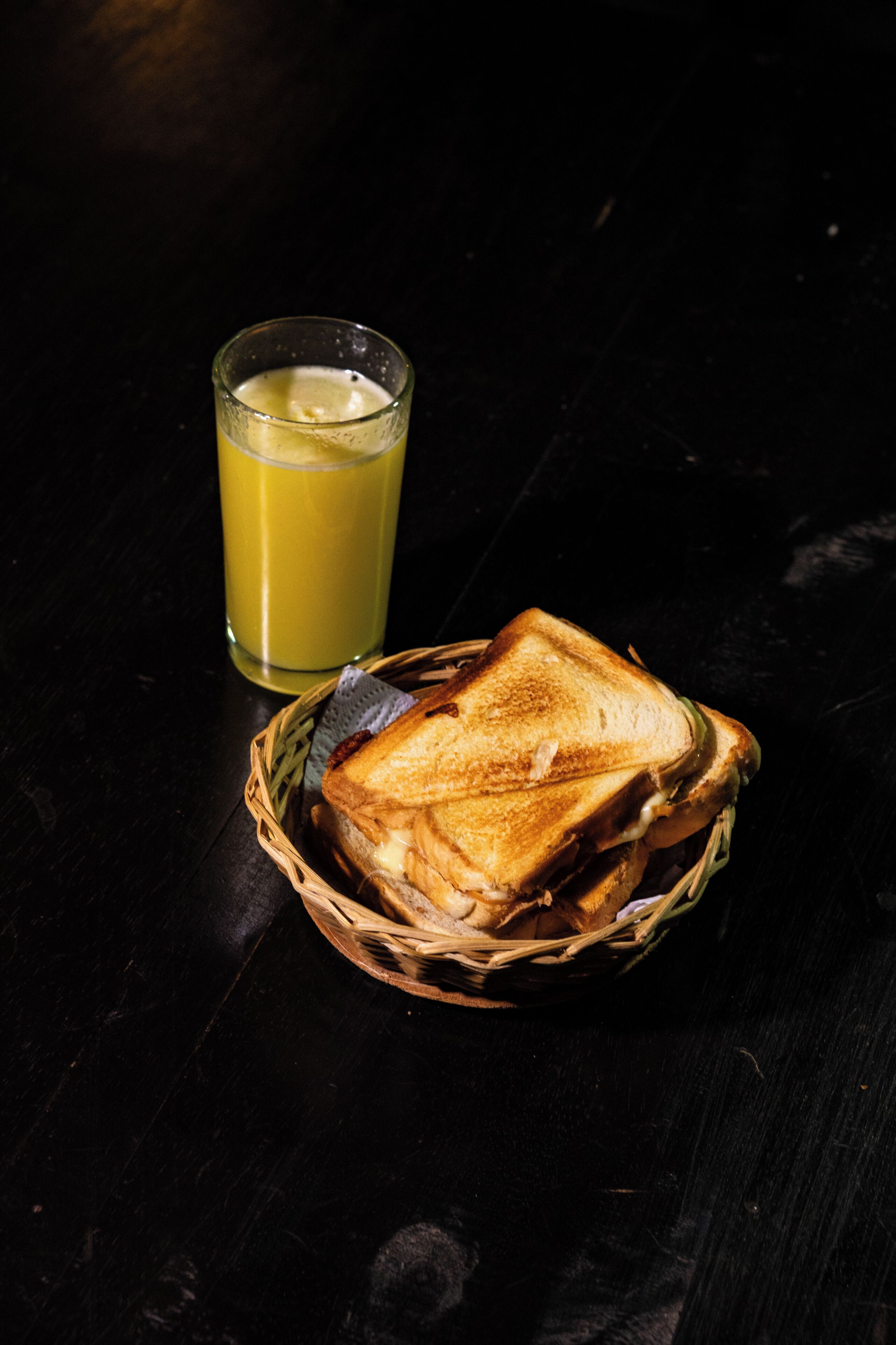 A sandwich in a basket and a glass of juice on a dark surface.