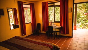 Una habitación con cama, un escritorio y sillas, una ventana con cortinas y vistas al verdor exterior.