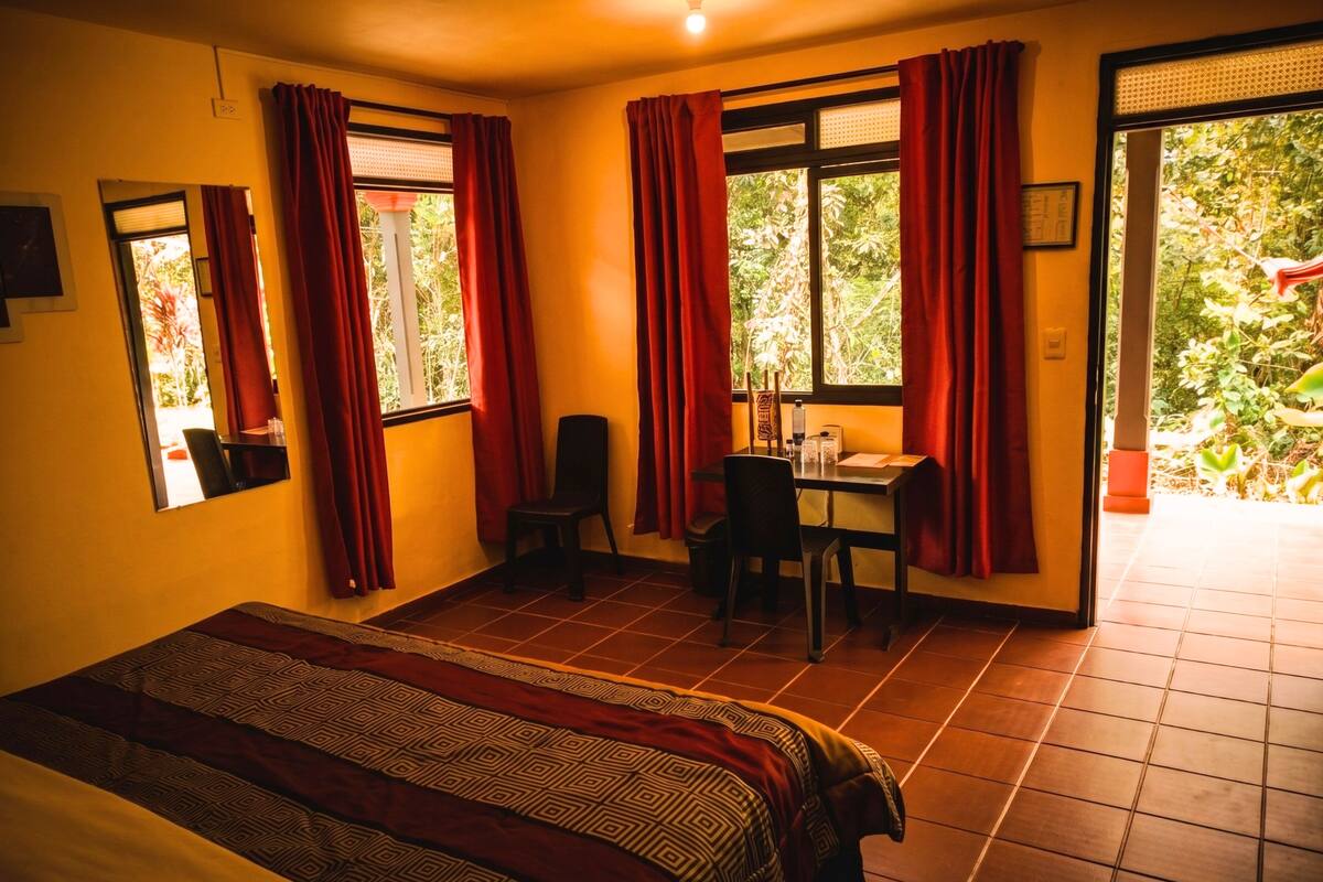 Una habitación con cama, un escritorio y sillas, una ventana con cortinas y vistas al verdor exterior.