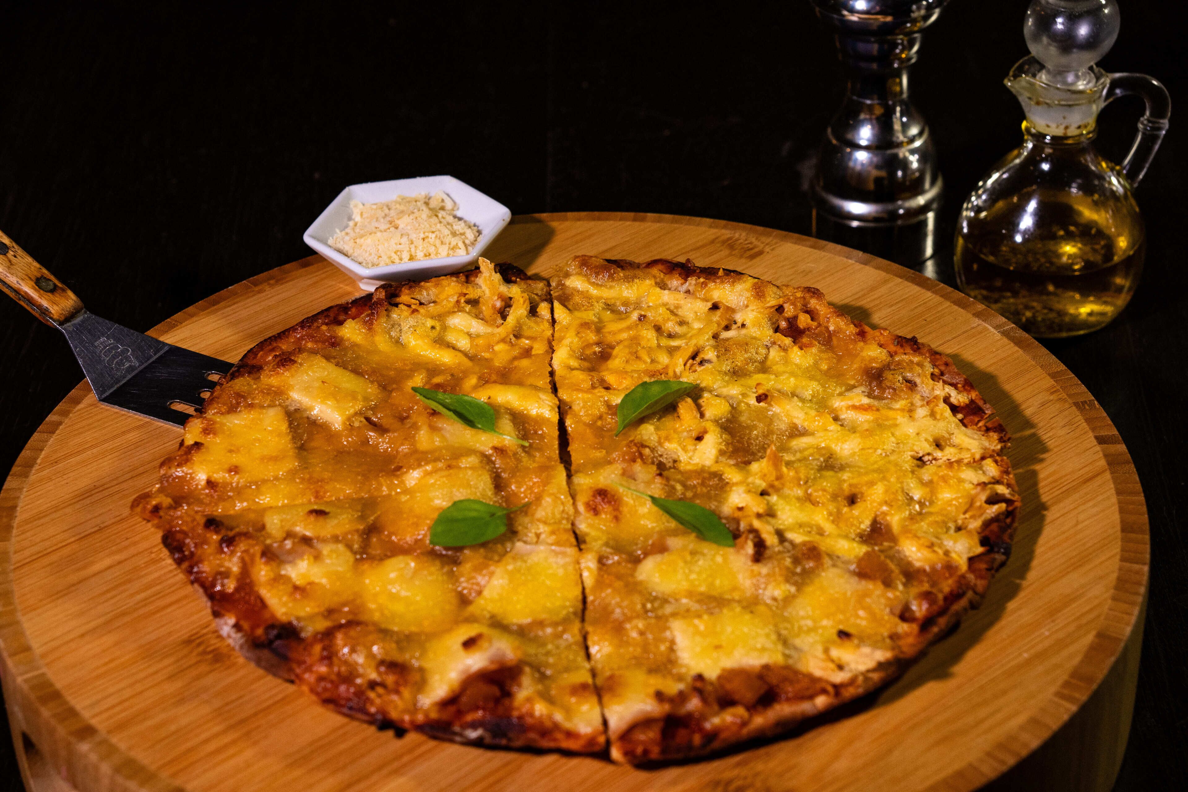 A pizza on a wooden board with a pizza cutter and a small bowl of seasoning.