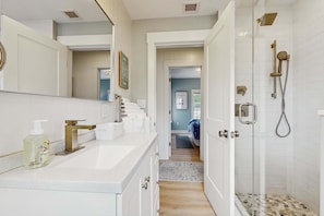 Un baño con un espejo grande, un lavabo, una ducha y una puerta que da a un dormitorio.