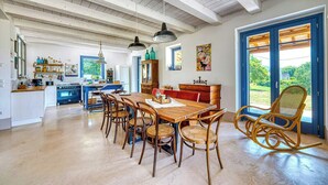 Une cuisine spacieuse avec des poutres en bois, un coin repas avec des chaises en osier et une vue dégagée à travers de grandes portes aux montants bleus.