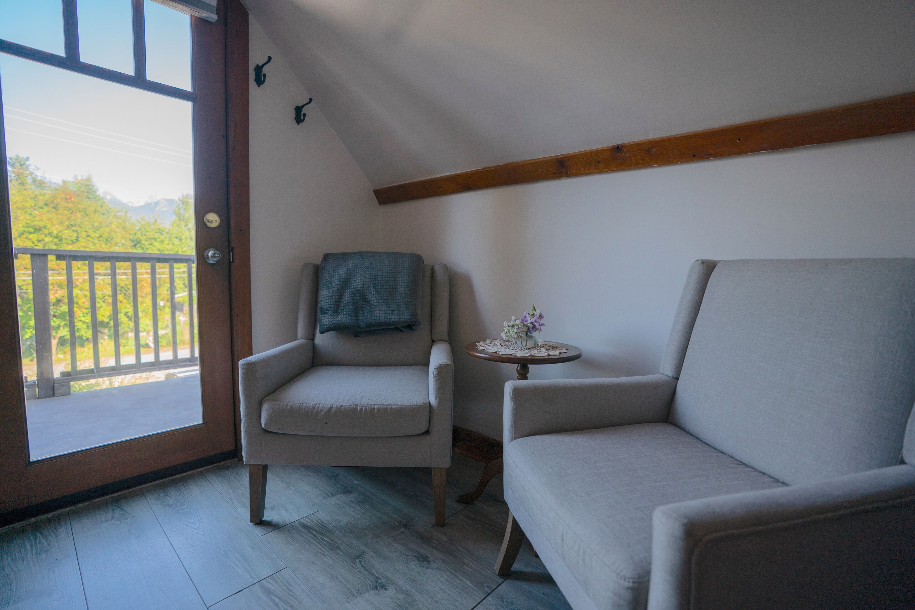 A room with a wooden door leading to a balcony, a small round table with a vase of flowers, and two armchairs.