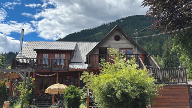A two-story wooden house with a balcony, a thatched umbrella, and a mountain backdrop. - Kaslo Manor Lodge - The Garden Suite (Kaslo)