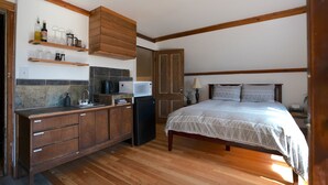 A compact bedroom with a bed, a nightstand, and a dresser. A kitchenette is visible with a refrigerator, microwave, and shelves. - Kaslo Manor Lodge - The Garden Suite (Kaslo)