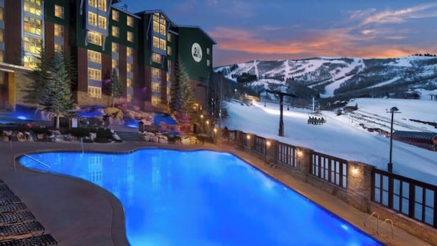 Um hotel com piscina, montanhas cobertas de neve e um teleférico ao fundo.