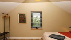 Une chambre avec un plafond en pente, un lit recouvert d’une couvre-lit à motifs, une fenêtre avec des stores, un tableau abstrait encadré et un porte-vêtements suspendu.
