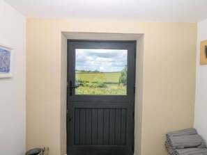 A door with a view of a green field, a framed picture on the wall, and a metal bucket on the floor. - Reddaway Byre (Okehampton)