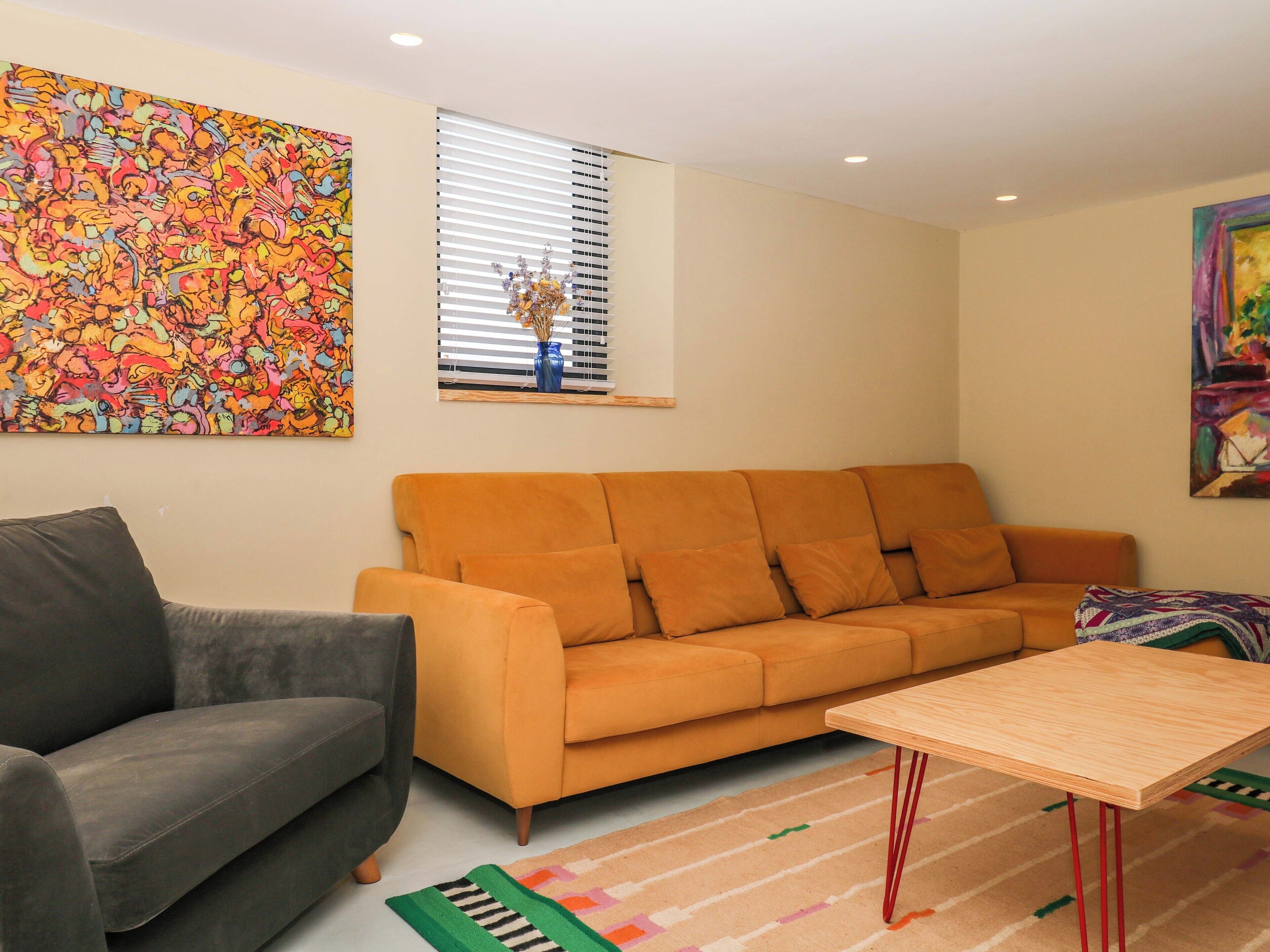 Una sala de estar con un sofá gris, un sofá naranja, una mesa de centro de madera y coloridas obras de arte en las paredes.