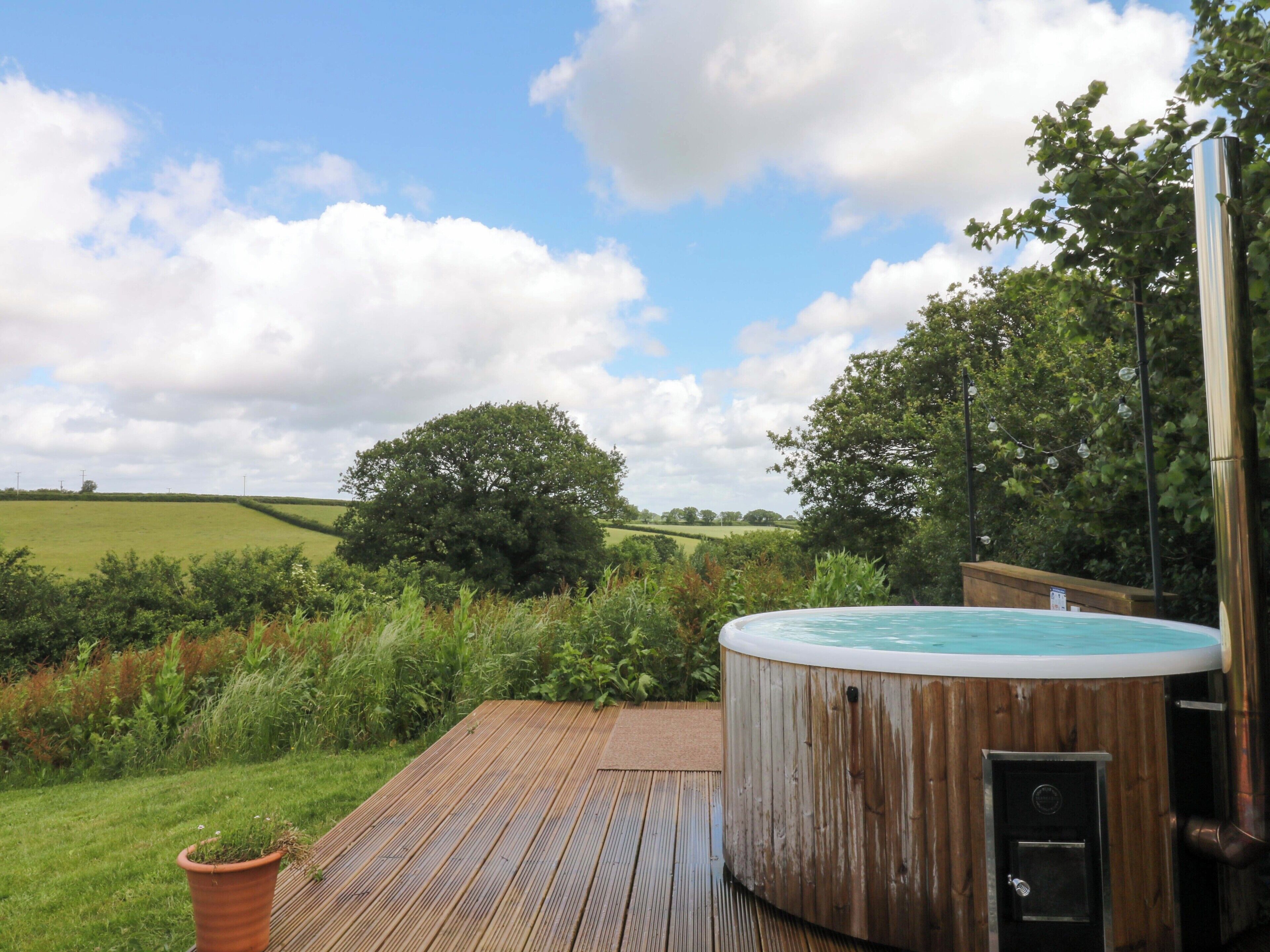 A hot tub on a wooden deck with a view of a green field and trees under a partly cloudy sky.