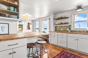 A kitchen with a wooden island, bar stools, white cabinets, and a view of the outdoors. - Island View Cottage - NEW! Waterfront Home in LDI (Little Deer Isle)