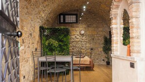 Une pièce aux murs de pierre, avec un lit, une table entourée de chaises et une horloge au mur.