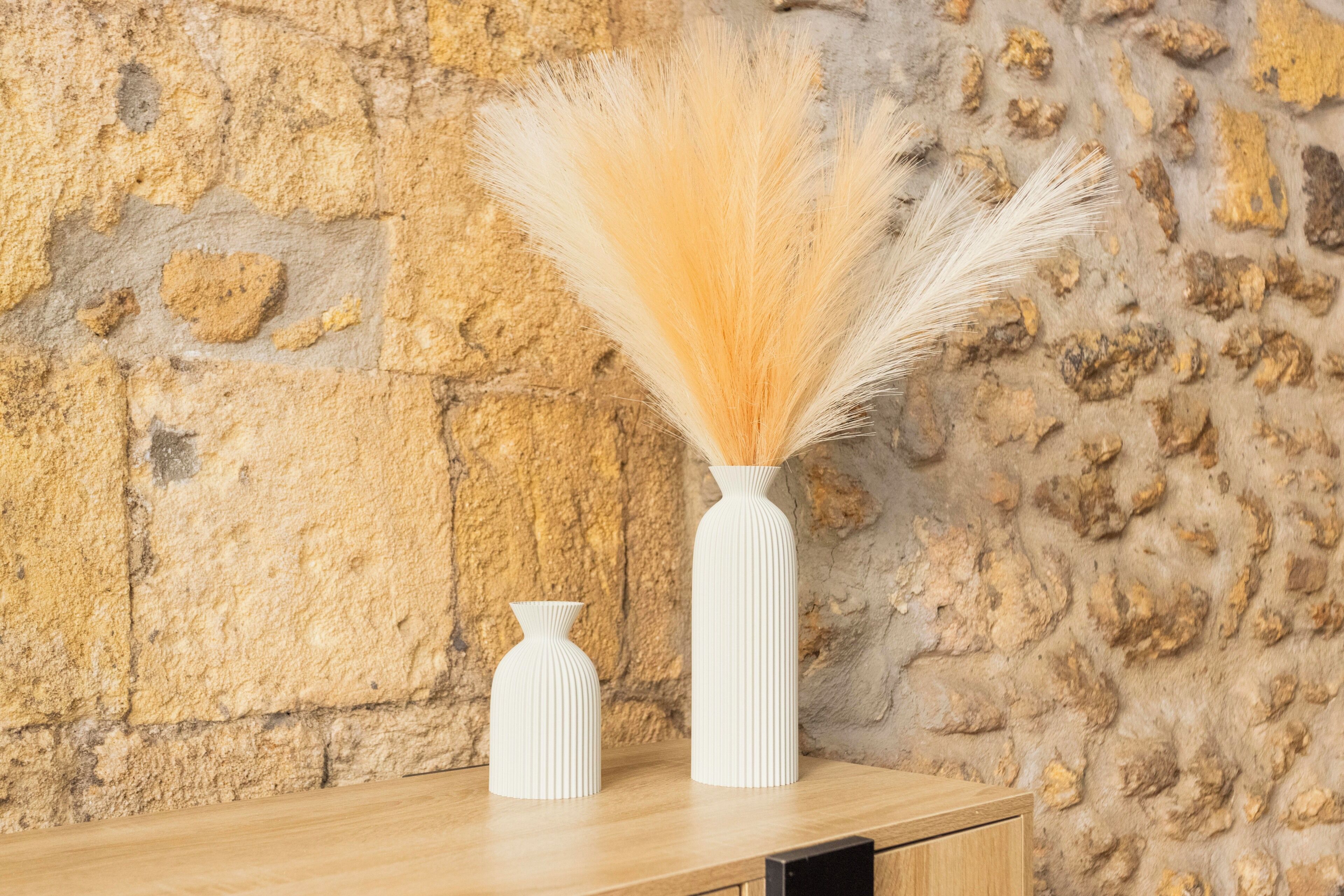 Two white vases with tall, light-colored grasses on a wooden surface against a textured stone wall.