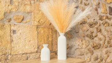 Two white vases with tall, light-colored grasses on a wooden surface against a textured stone wall.