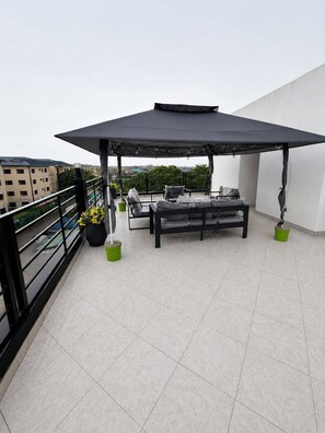 Eine Dachterrasse mit überdachter Sitzecke, Kübelpflanzen und Blick auf Gebäude im Hintergrund.