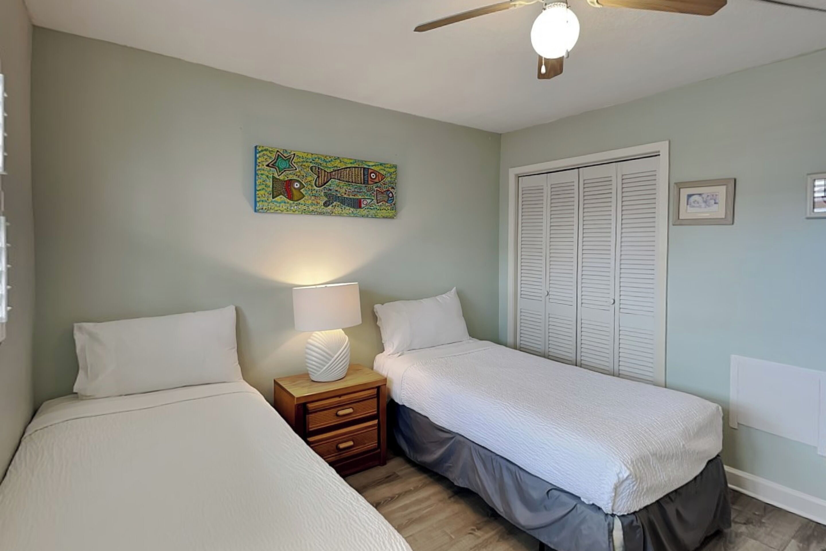 A bedroom with two beds, a nightstand, a ceiling fan, a wall-mounted artwork, and a white door with shutters.