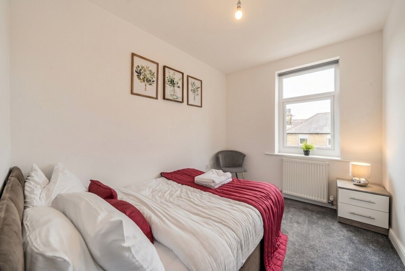 Ein Schlafzimmer mit einem Bett, einem Sessel, einem Nachttisch mit Lampe, einem Fenster und drei gerahmten Bildern an der Wand.