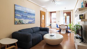 Ein Wohnzimmer mit Sofa, Couchtisch und Sesseln, einem Gemälde an der Wand und einem Fernseher rechts.