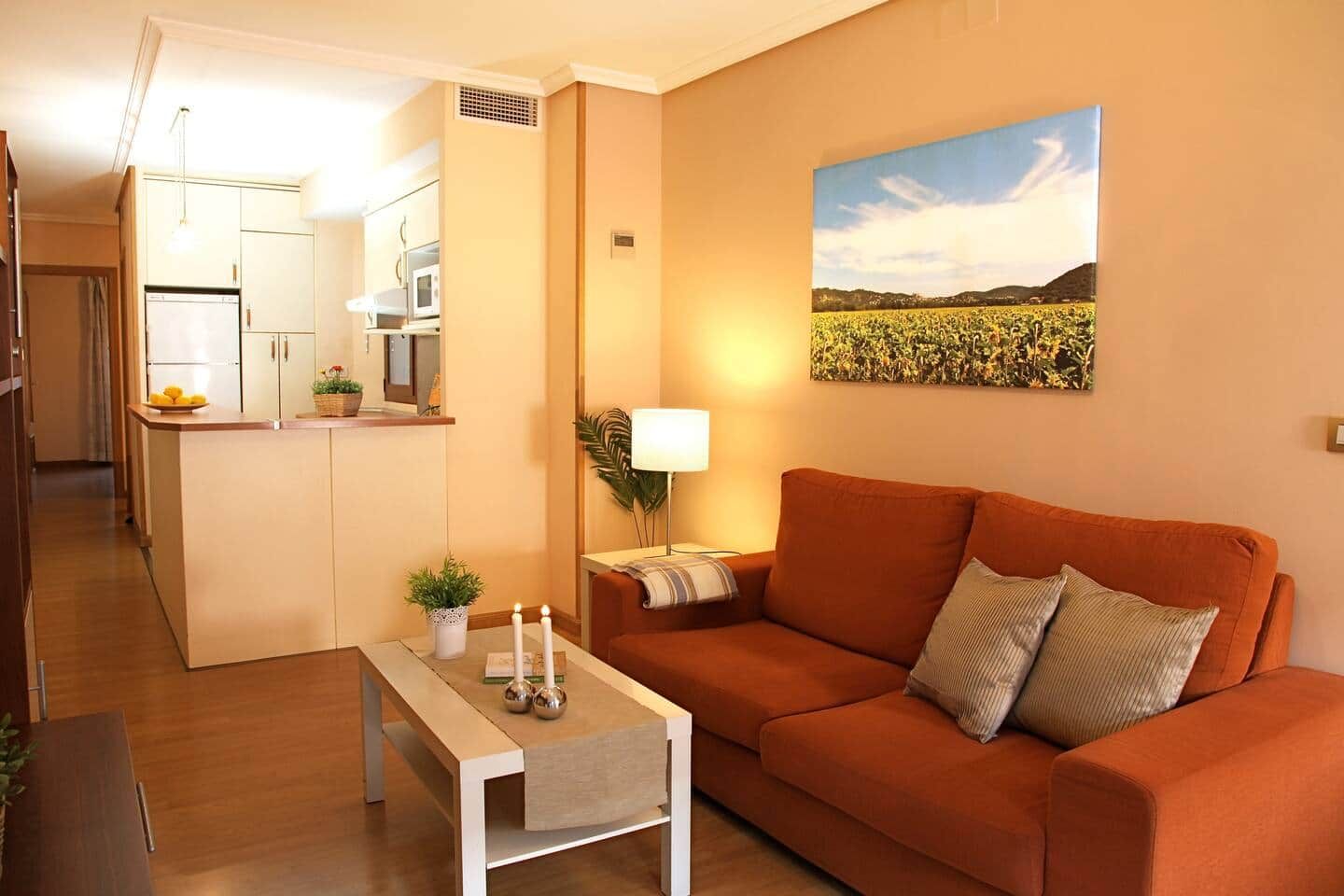 Una sala de estar con sofá, una mesa, una lámpara y una zona de cocina con armarios y electrodomésticos.