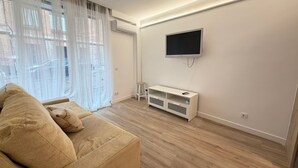 Un salon avec un canapé, un téléviseur fixé au mur et une fenêtre ornée de rideaux légers.