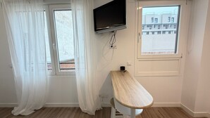 Une pièce avec deux fenêtres, un téléviseur fixé au mur et un tabouret en bois.