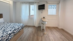 Une chambre à coucher avec un lit, un téléviseur fixé au mur, une fenêtre avec des rideaux et un petit tabouret.