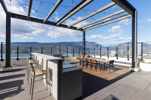 A rooftop terrace with a bar area, seating, and a view of the city and mountains.