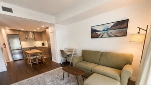 Un salon moderne avec un canapé, un pouf et une table à manger avec des chaises. On aperçoit au fond une cuisine équipée d’un réfrigérateur et de placards.