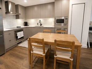 A modern kitchen with a wooden dining table and chairs, a built-in oven, a microwave, and a stainless steel range hood.
