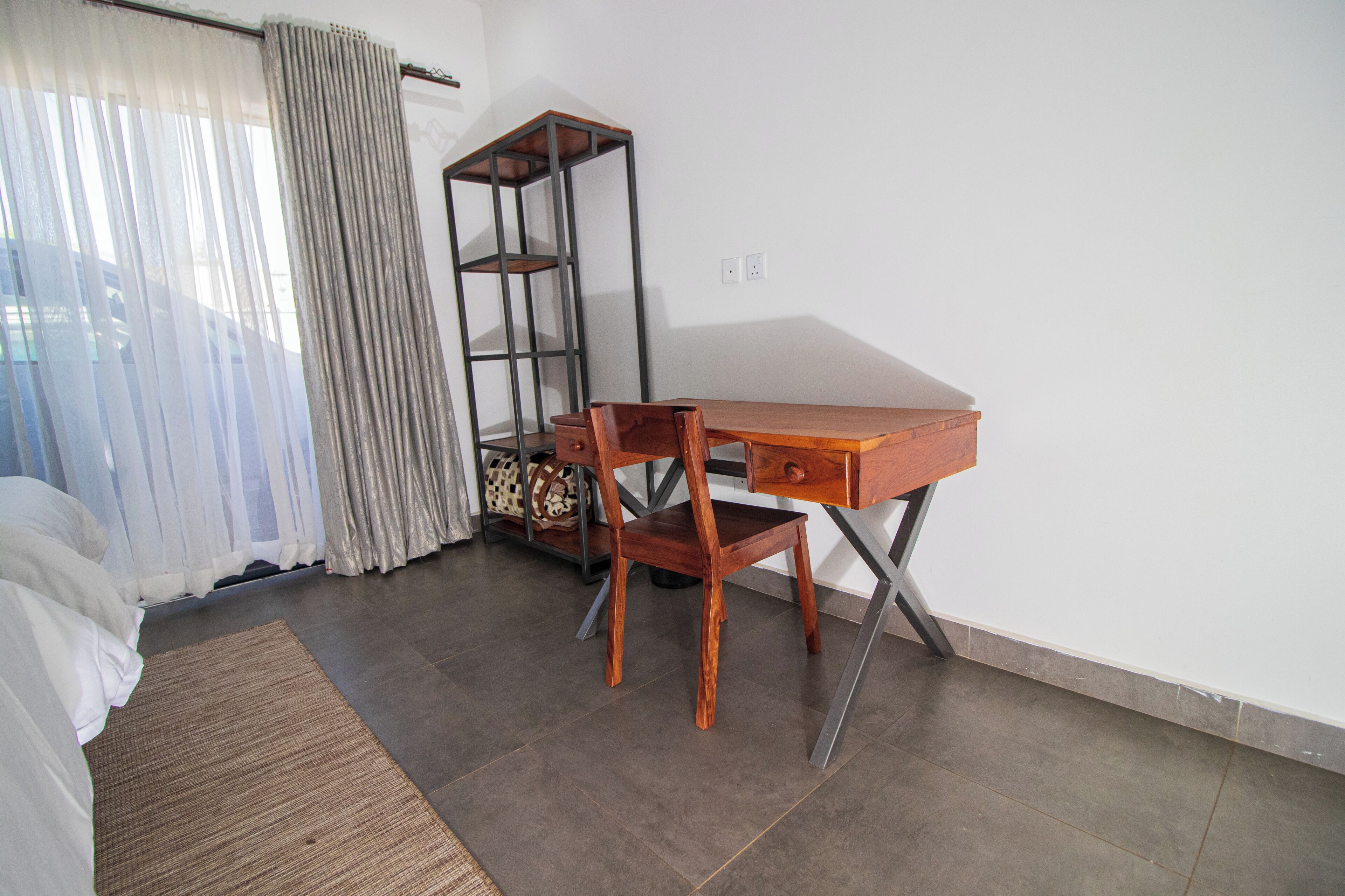 A room with a bed, a wooden desk, a chair, a shelf unit, and a window with curtains.