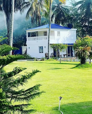 Un bâtiment blanc à deux étages, surmonté d’un toit bleu, entouré d’une végétation luxuriante et d’une pelouse bien entretenue.