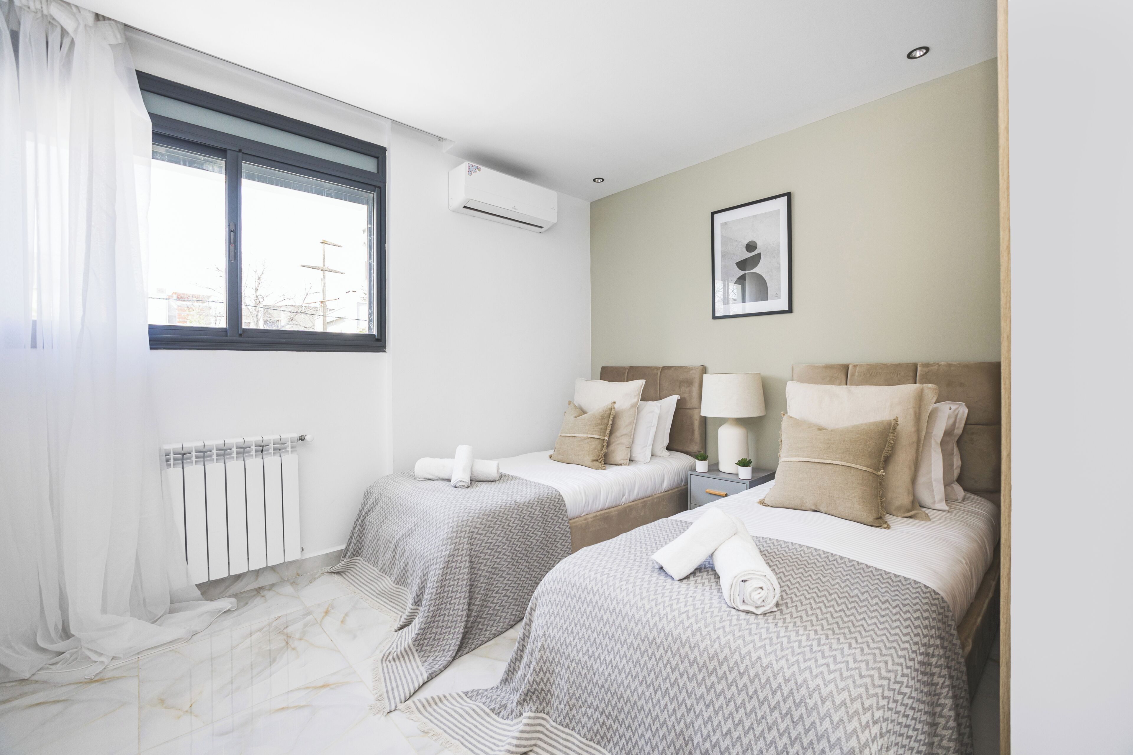 A hotel room with two beds, a window with curtains, an air conditioning unit, a framed picture on the wall, and a bedside table with a lamp.
