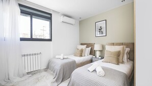 A hotel room with two beds, a window with curtains, an air conditioning unit, a framed picture on the wall, and a bedside table with a lamp.