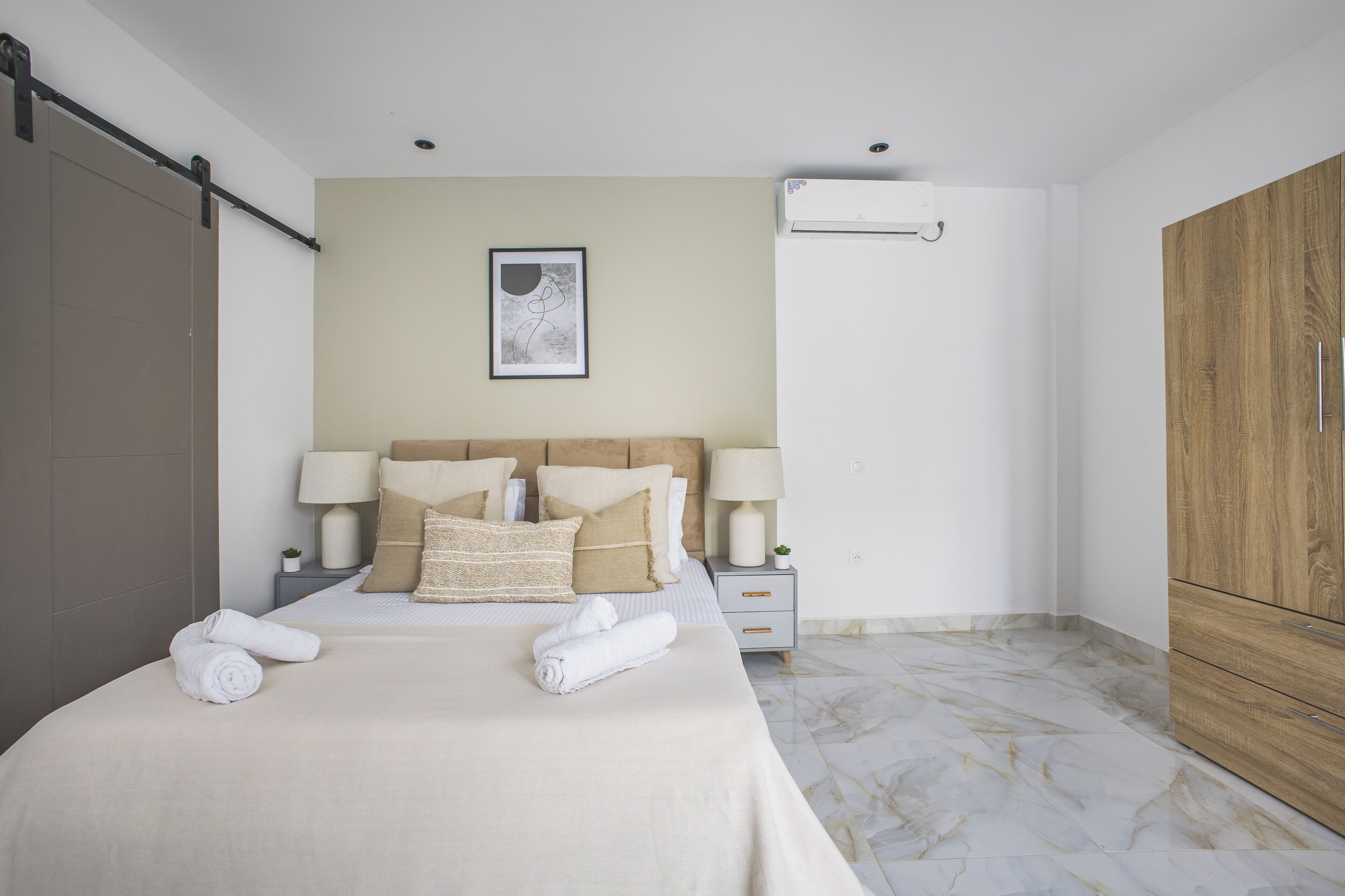 A neatly made bed with pillows and two rolled towels, a bedside table with a lamp, a wall-mounted air conditioner, and a wooden wardrobe.