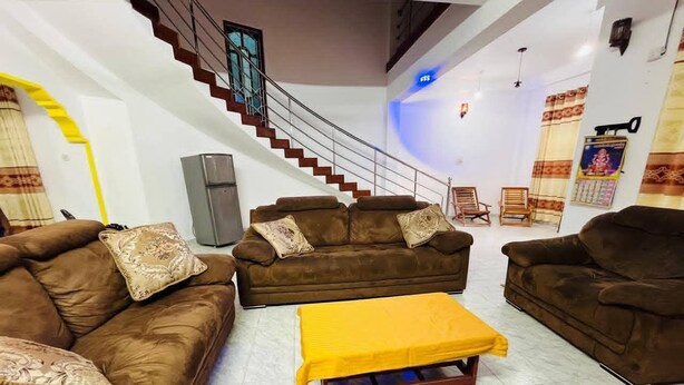 A living room with brown sofas, a yellow ottoman, a staircase, and a wall-mounted TV. - Kamaleswari Villa Trincomalee | 3BR House | 2km to Beach | Family Stay (Trincomalee)