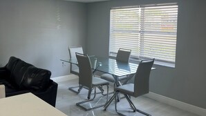 Una sala de reuniones moderna con una mesa de vidrio, sillas metálicas, un escritorio blanco y una ventana con persianas.